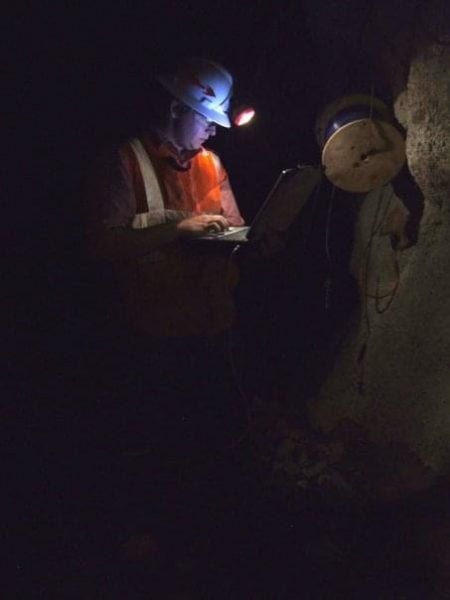 Walt is standing outdoors, focused as he downloads data from a small device attached to his headlamp, illuminated by soft light in the background.