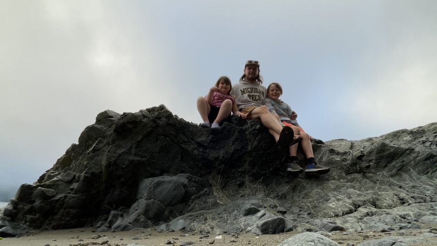 Walt and his kids are camping on the west coast of Vancouver Island in 2022, gathered around their campsite with a scenic coastal backdrop of trees and ocean.