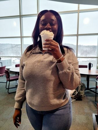 A woman, Angela, stands in front of large windows with a view of the outdoors, holding a steaming cup of hot cocoa in both hands. She is gazing contentedly, with soft natural light highlighting her face and casting gentle shadows behind her.