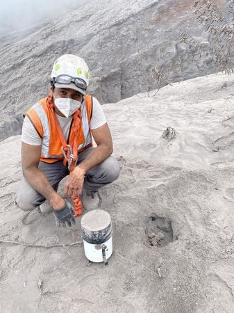 On the steep slope of a volcano, Gustavo Bejar-Lopez wears a helmet and respirator mask while crouching to collect volcanic material samples in rocky, ash-covered terrain.
