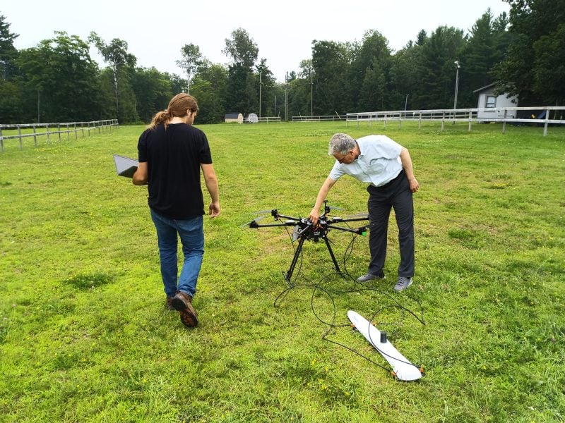 Sam Jensen and Aleksey Smirnov prepare a drone for its first outdoor flight, with tall trees in the background.