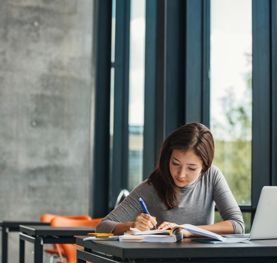 Young woman reading a book and taking notes at a desk, in front of a window in an open online setting. This image demonstrates that online learning  can happen anywhere.