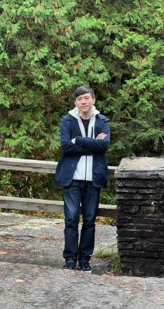 Yipeng leaning against stone pillar in front of evergreen trees