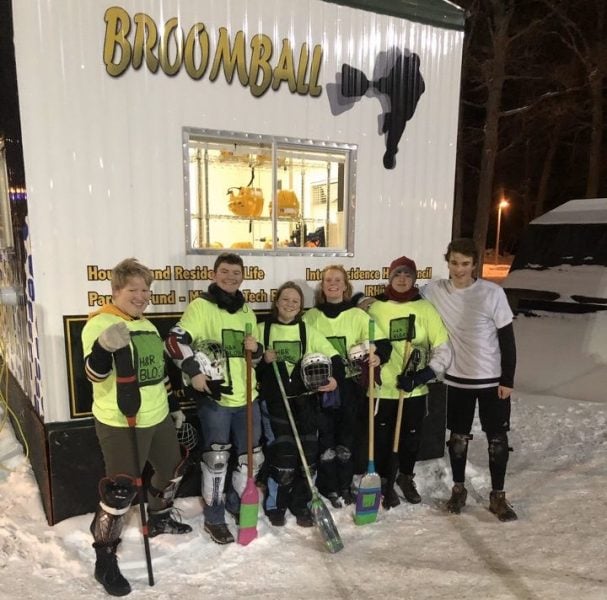 Students in matching jerseys hold broomball sticks in front of a food trailer