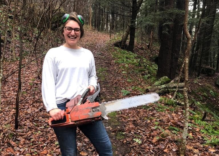A young woman laughs in the woods, wearing ear protection and brandishing a chainsaw.