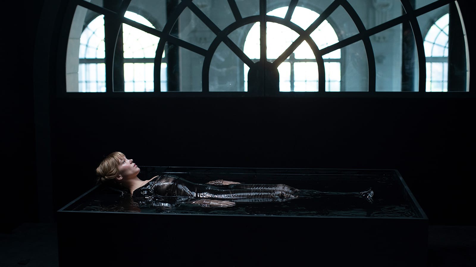 An attractive young woman lays in a rectangular black tub filled with black liquid below a large half-circle window with washed-out white light shining through.