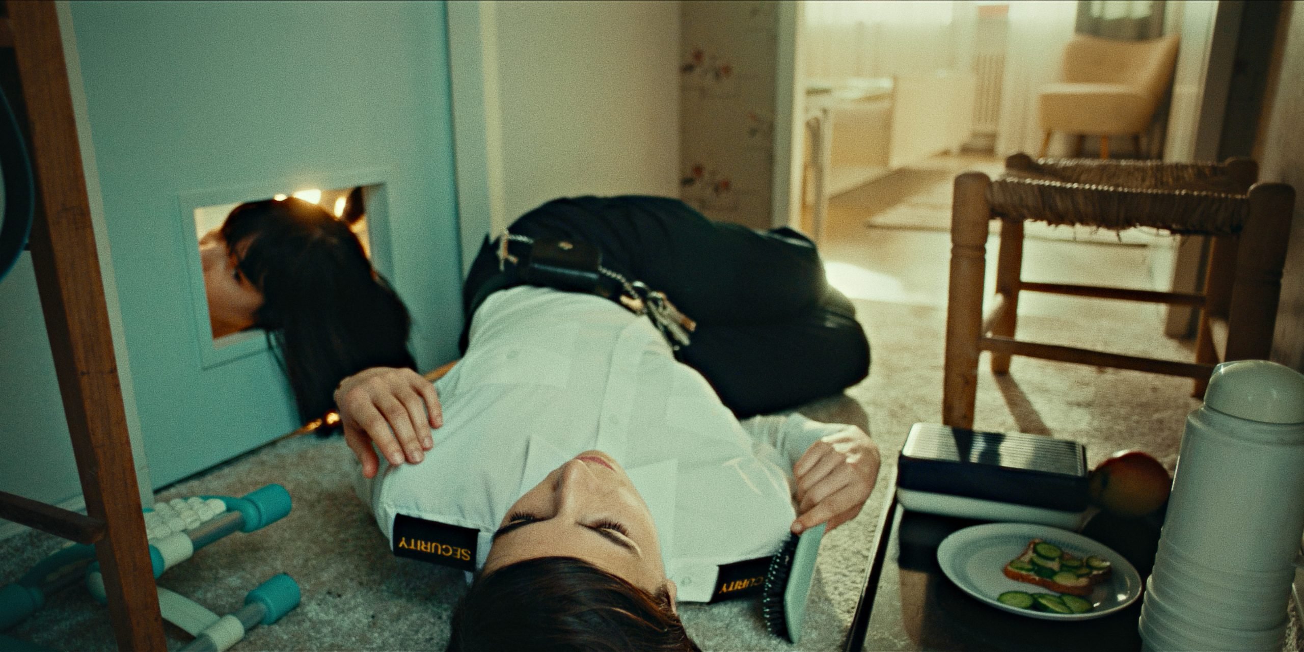 Still from "We Might As Well Be Dead showing a woman in a security uniform laying on the floor of a cramped apartment. Next to her another woman's head can be seen through the mail slot of a closed door with her hair hanging through.