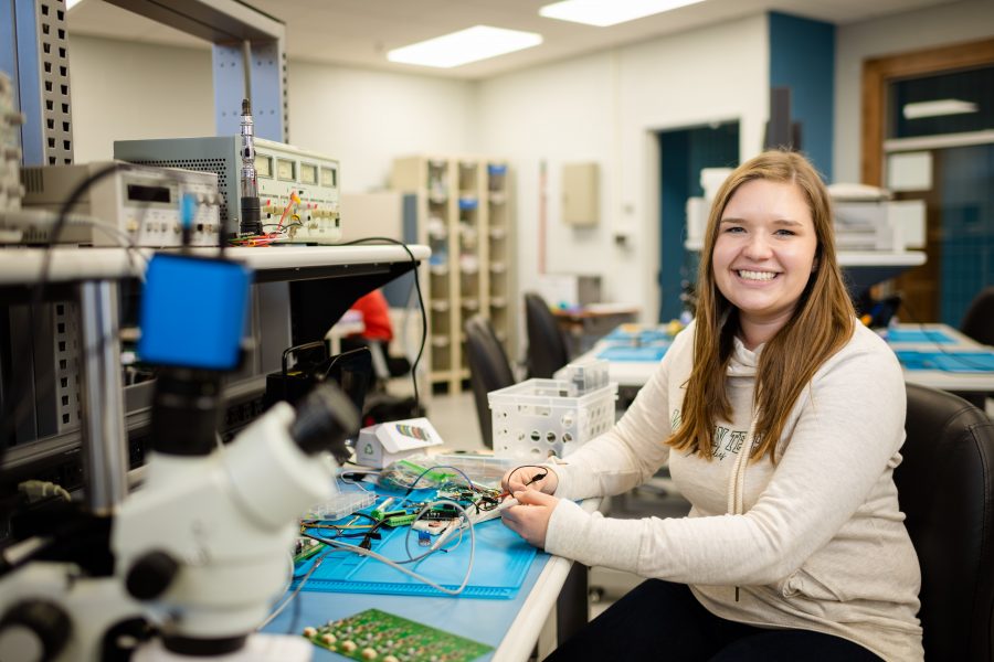 A young woman wearing a Michigan Tech shirt sits at a circuit board in an electronics makerspace at Michigan Tech with electronic equipment behind her.