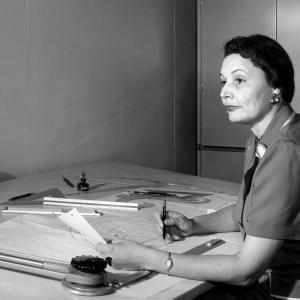 Mary Golda Ross sits at a drafting table holding a pen.