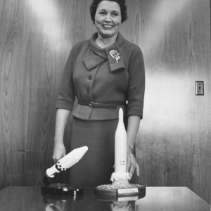 Mary Golda Ross stands smiling before a table with two models of spacecraft she worked to design.