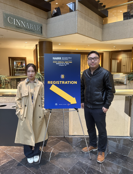 Xiaoyu Hou and Dr. Bo Xiao stand next to a NAIRR Conference Registration poster in a hotel lobby.
