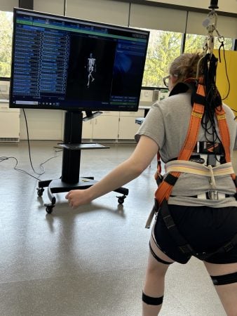 Study subject hooked up to a harness with a number of sensors attached to their body facing a large screen showing their body movement and movement statistics