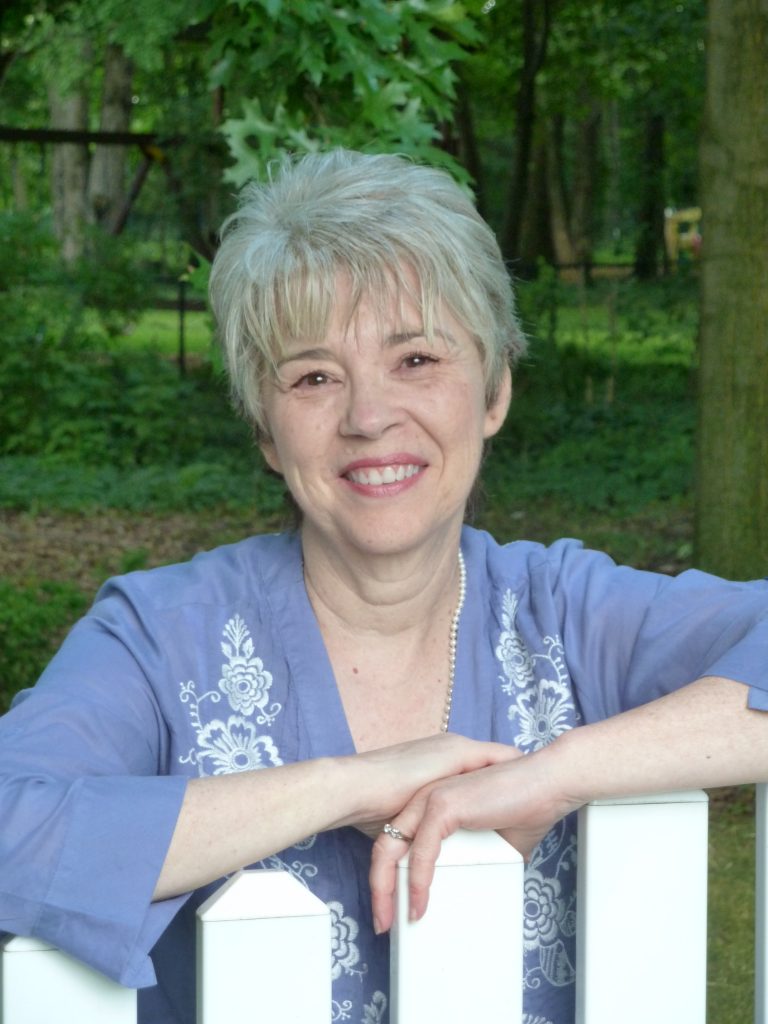 Publicity photograph of author Mary Doria Russell. A woman poses and smiles by a fence.