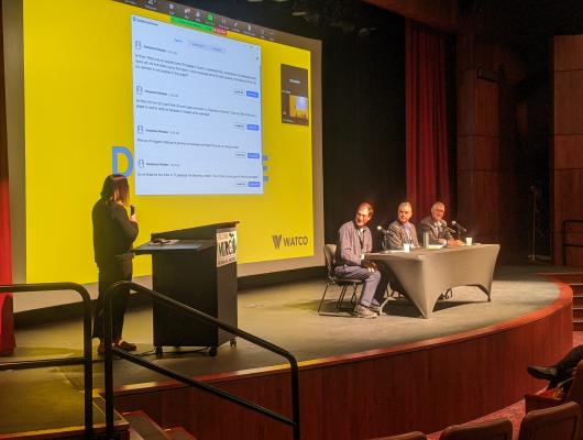 A panel and speaker on stage with a presentation on the screen.
