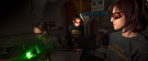 Three biomedical engineering students work in a darkened Biomedical Optics Lab with laser in safety glasses.