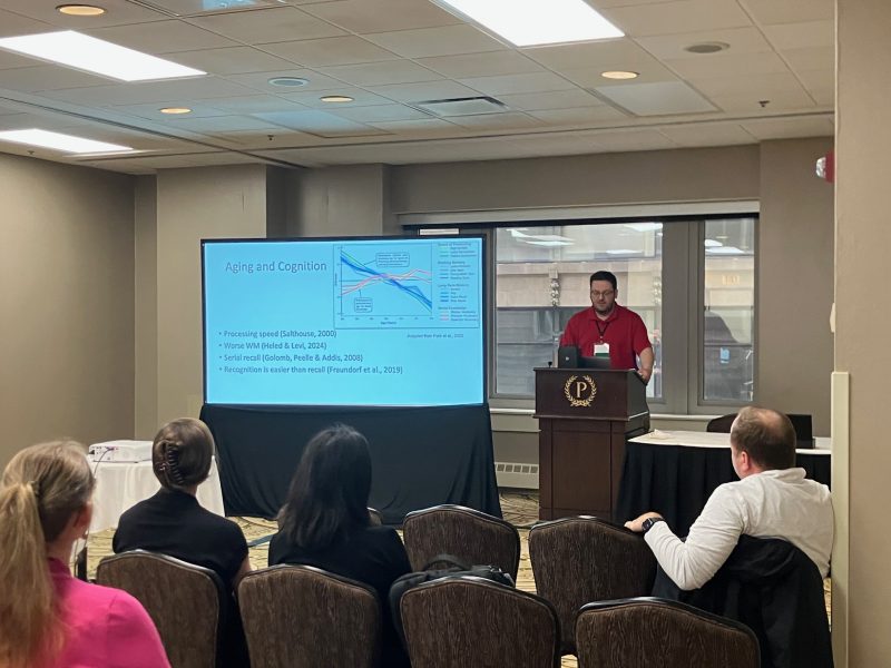 Brandon Woolman standing at a podium in front of a crowd presenting his research “Characterizing Age Difference in a Novel Motor Sequence Learning Task.” 
