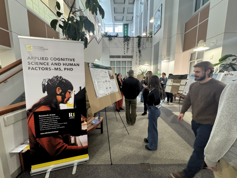 MTU and NMU students mingle around easels displaying research posters in a hall at NMU