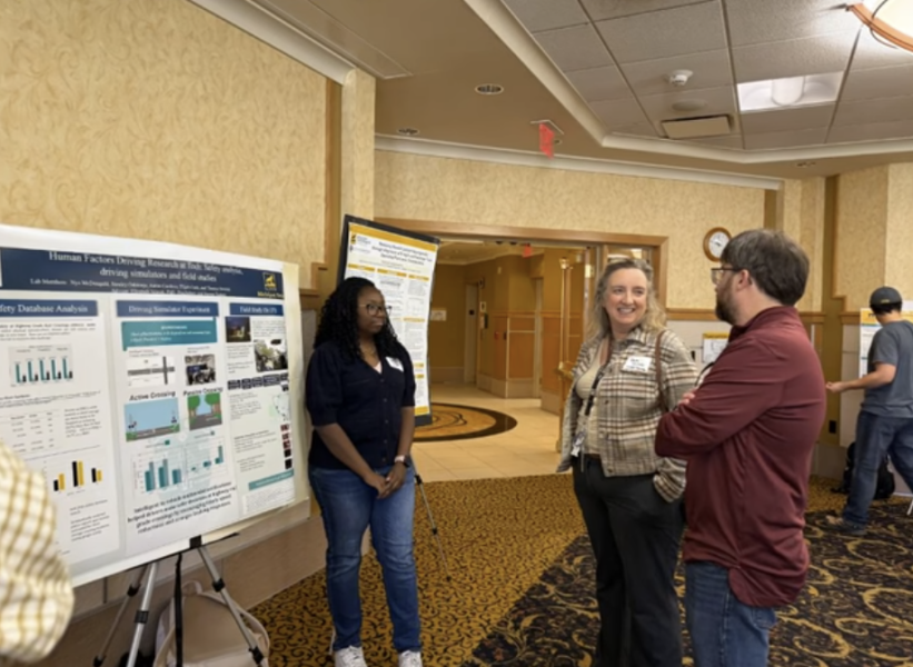 A young woman stands near a research poster while her professor, also a woman, stands near by 