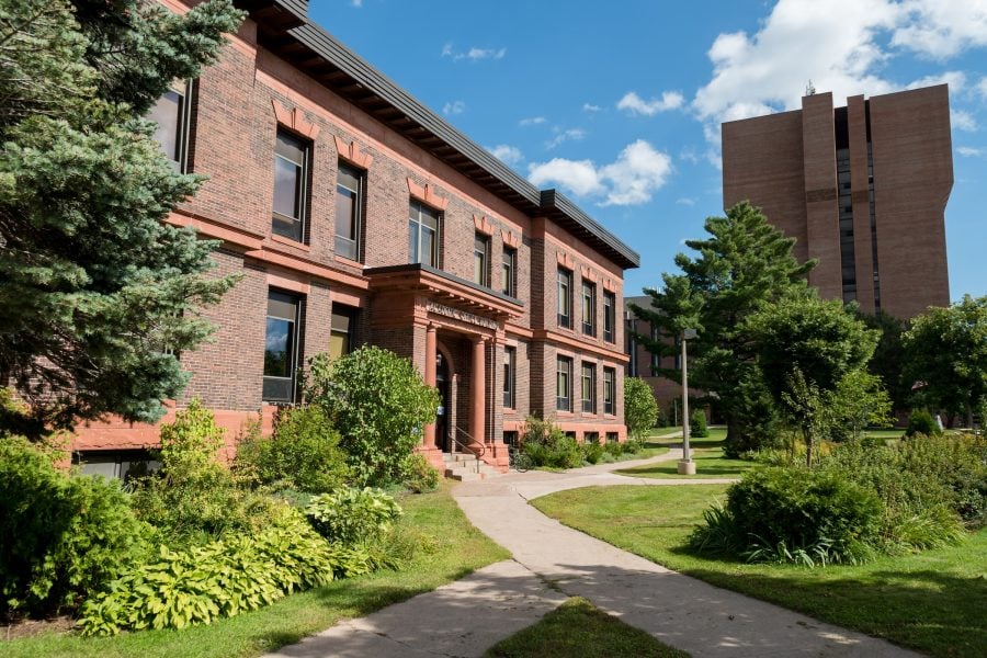 Brick two story building with concrete walkways in front, surrounded by green grass, shrubs, and trees.