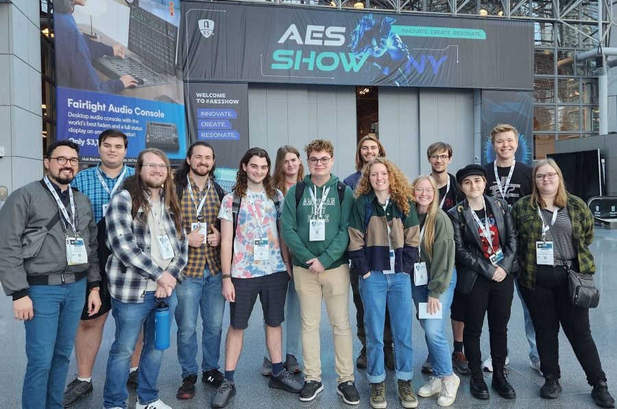 Thirteen students stand with assistant professor Jeff Sherwood in front of a large hanging AES Show banner at the student recording competition.