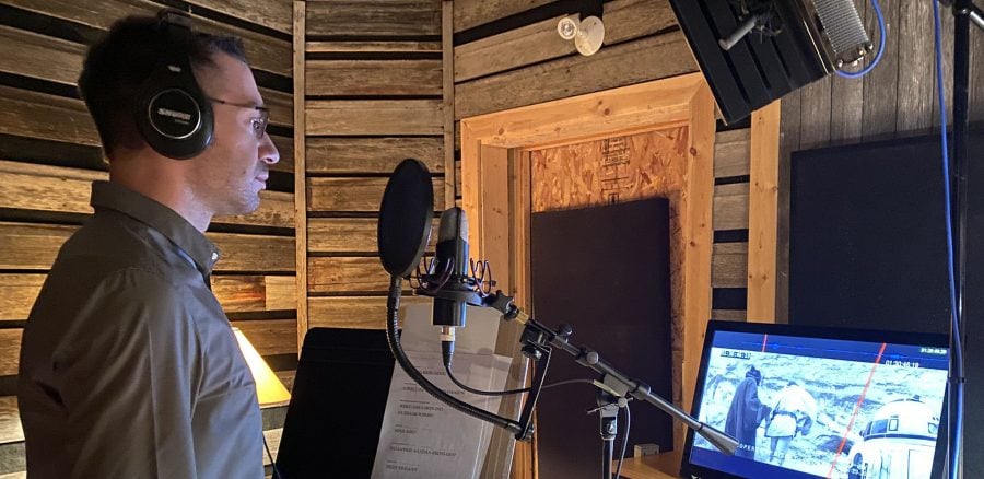 Kalvin Hartwig stands in a sound-proofed recording studio in front of a computer, microphone and recording equipment as he records lines for his voice acting role.