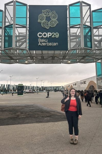 Lexi Tater stands in front of an exterior glass arch, a sign which reads, “COP29 Baku Azerbaijan.”