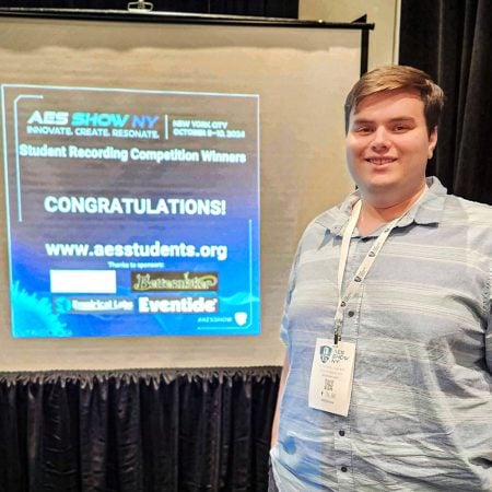 Nate Lyons stands in front of a projector screen at the AES conference. The message on the screen congratulates him on winning the Student Recording competition.