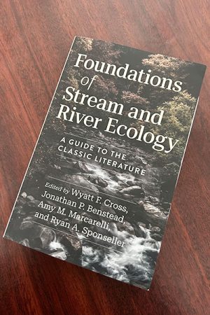 The book sits on a brown table. The cover art shows a rocky stream in the forest. The text reads: “Foundations of Stream and River Ecology: A Guide to the Classic Literature Edited by Wyatt F. Cross, Jonathan P. Benstead, Amy M. Marcarelli, and Ryan A. Sponseller
##