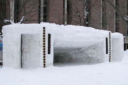 A snow structure roughly the size of two vehicles sits in front of Michigan Tech’s R. L. Smith Building. It has three rectangular, cone-like segments divided by two columns of speakers.