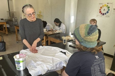 Anne Beffel talks with student Evan Meyer in an art classroom. A plain white t-shirt sits on the table between them.