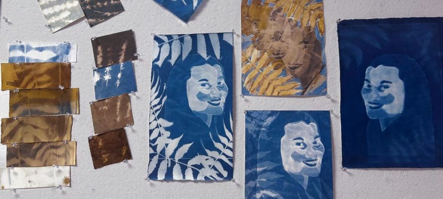 ultiple Cyanotypes stained and painted with natural inks hand together on a white wall. Four on the right depict variations of a woman’s face with leaf patterns. Several on the left depict leaf and flower patterns on their own.