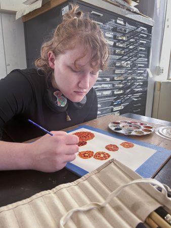 Mallory Grant sits at an art classroom table painting chrysanthemums with orange, red and yellow watercolors.