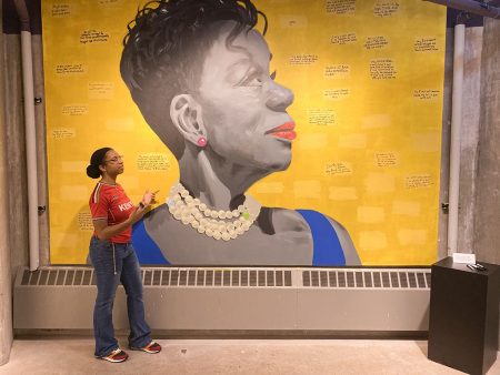 Allison Lewis stands in front of a large mural, gesturing to the painting behind her. The background of the painting is a bright yellow sprinkled with personal stories and names written by visitors in black permanent marker. In the center of the mural is a profile of a woman from the shoulders up. She is looking up and to the right off the canvas. The woman has short hair, and is in grayscale aside from her red lips, pink earrings, white pearl necklace and blue shirt.