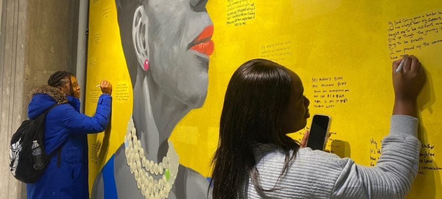 Two people stand writing on a large mural in black permanent marker. The mural has a bright yellow background, with a desaturated painting of a woman’s shoulders and face. Red lipstick, pink earrings, white pearls and a blue shirt stand out brightly against her grayscale skin tones and hair. There are many other writings visible on the yellow background surrounding the painted woman.
Allison Lewis and Terri Frew.