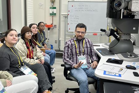 Conference attendees sit along one wall in a lab while a workshop leader discusses the microscope and other physics equipment in front of them.