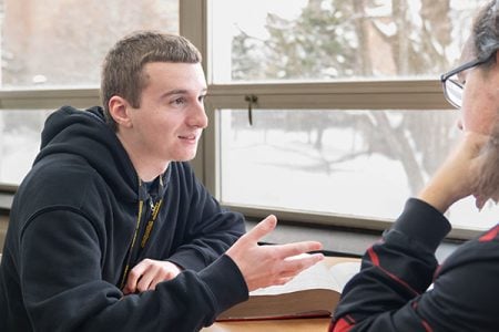 McCoy Ziehl sits at a study table in front of a window, talking and gesturing to another student. A book sits open on the table between them.