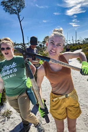 hree students work on a beach. Skyler walks towards the camera with her hand on her water bottle, mouth open and smiling. Ben stands behind her, also smiling. Olivia poses in the center of the photo with a large set of pruning shears, pulling a comical face.