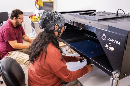 An undergraduate student sits at a large black Kinarm machine, wearing a helmet-shaped device to track brain waves which another student is observing on a computer screen in the background.