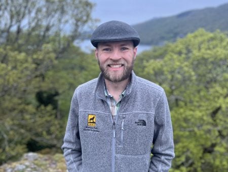 Mark Rhodes smiles at the camera, wearing a zip up Michigan Tech branded fleece in front of a scenic view of trees, hills and a lake on a cloudy day.