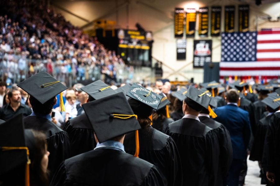 A large group of students in graduation caps and gowns walk away from the camera, toward the graduation podium at the other end of a large multipurpose hall. One graduation cap includes the decorative phrase, “Journey Before Destination.”