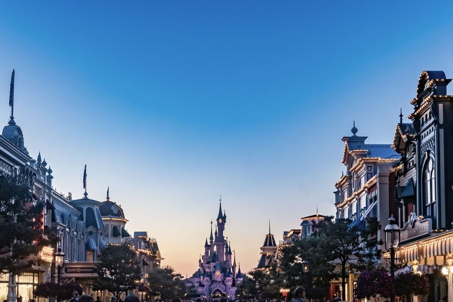 A photo of a street in Disney World’s Magic Kingdom, with Cinderella’s castle haloed by the pink glow of a setting sun in the background and the park’s store and attraction lights twinkling in the foreground under a blue sky.