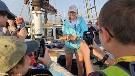 SYP students on a boat gather around a smiling instructor holding a live fish, eager to get a photo of the wriggling wildlife.