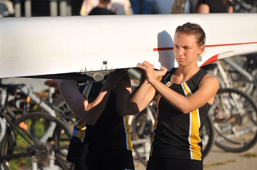 Aili Toyli, wearing her black and gold rowing team uniform, helps carry the boat with her teammates. She has an expression of pure determination.