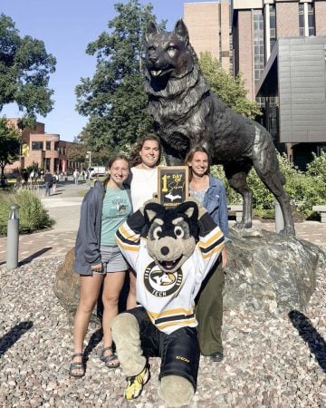 Aili Toyli poses in front of the rock dog statue on Michigan Tech’s campus with Blizzard and two other incoming students. Blizzard kneels in front of the students, holding a sign that says “1st Day of School #MichiganTech” with a gold image of the Upper Peninsula, facing the camera.