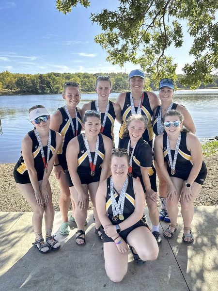 Aili Toyli and eight other rowing club members pose wearing winning medals on the shore of a body of water.