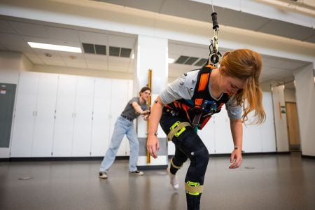 A participant in a safety fall harness leans forward in a lunge after allowing herself to fall while a researcher guides her through the process.