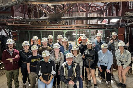 A large group of students and faculty wearing mining helmets in an industrial setting.