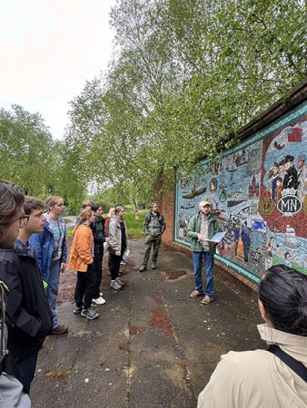 Mark Rhodes stands in front of a brick wall painted with a cultural mural, holding a binder and speaking to a group of students who listen attentively, facing the mural.