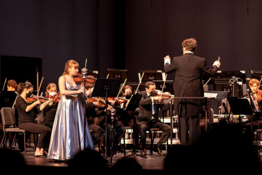 Joel Neves stands in front of an orchestra with his back to the camera, waving his baton.