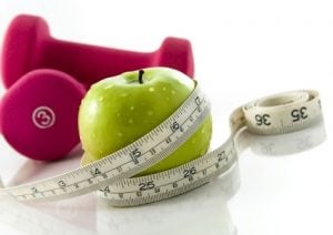 Fresh appetizing apple and brightly colored dumbbells tied with a measuring tape. Slight reflection, white background, focus on the apple